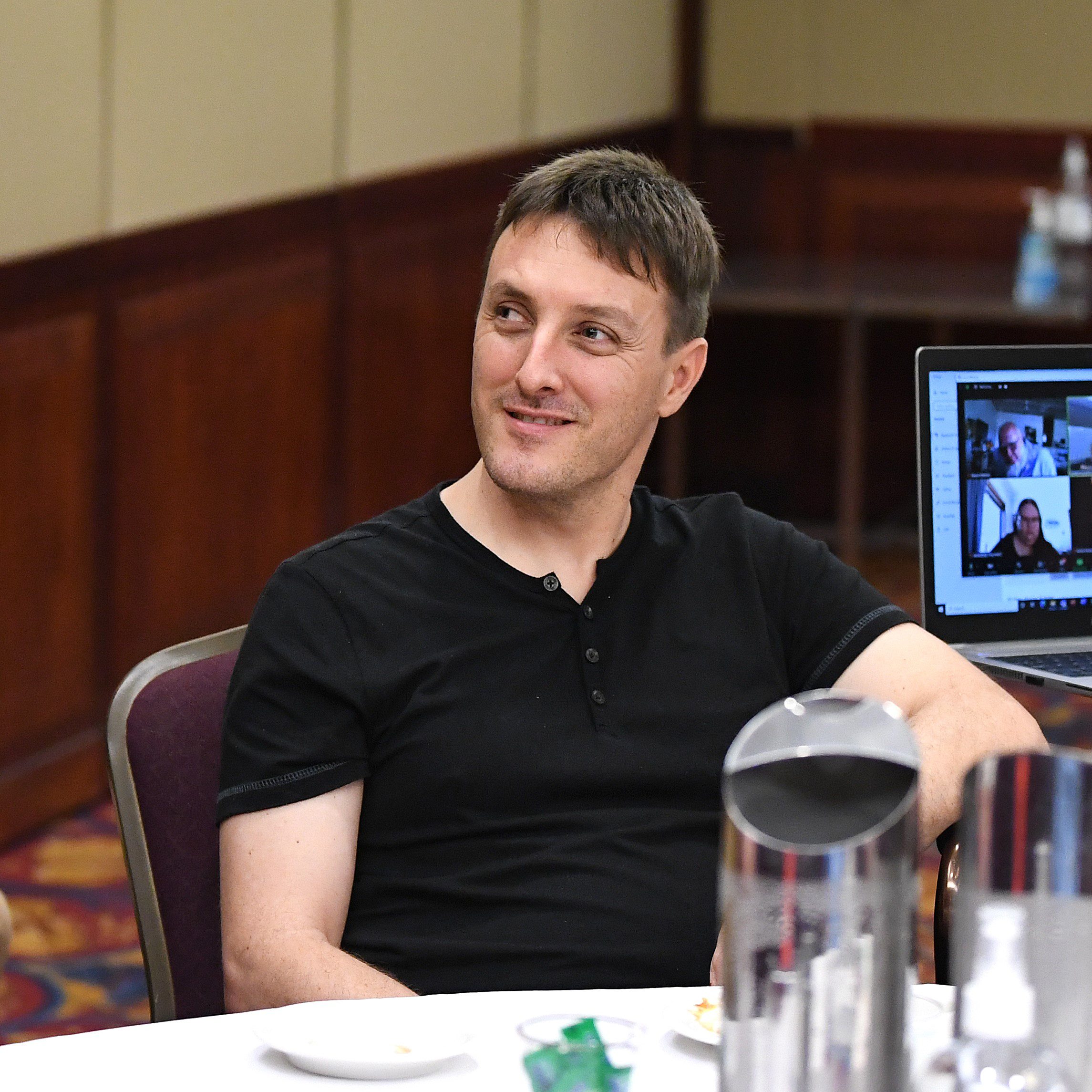 A man wearing a black shirt sitting at a table smiling at someone outside of the photo frame.