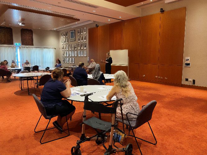 People are seated at multiple spread out tables in a large conference room. There is a lady standing at the front presenting to the group.