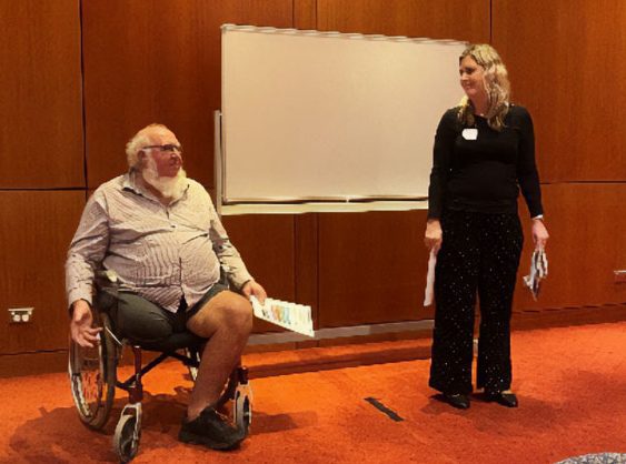 A man in a wheelchair is addressing people in a room and is next to a standing woman who is looking at him.