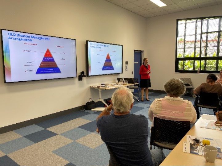 A lady is standing at the front of a room presenting to a group of four people sitting down. She is holding up a piece of paper and is standing in front of two projector screens. The content on the screens and paper are blurry.