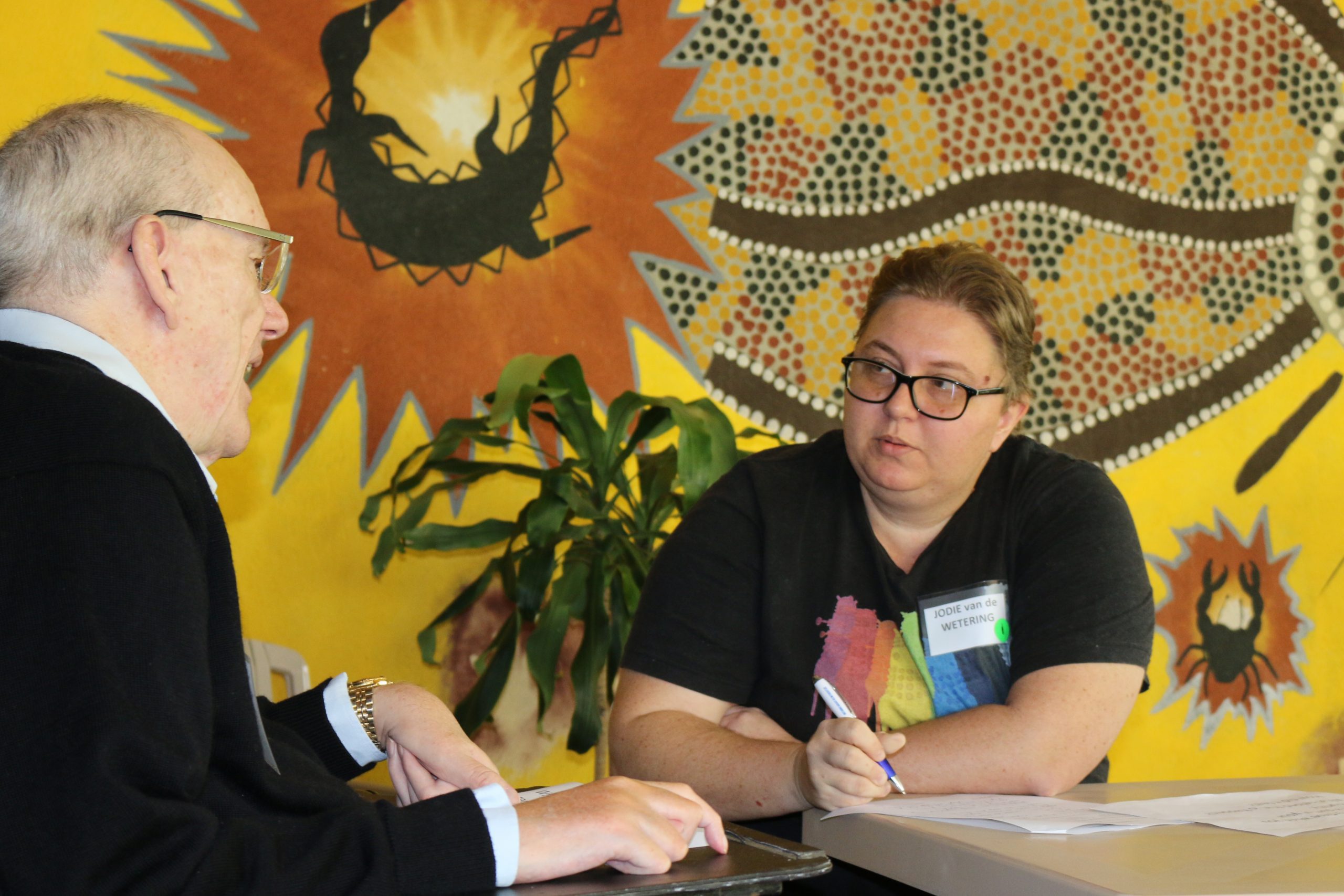 A man and a woman talking at a desk in front of a wall with Indigenous Australian art on it.