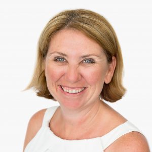 A woman with shoulder length blonde hair, wearing a white singlet top and smiling at the camera.