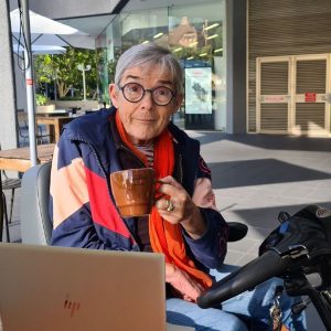 A lady sitting on an electric scooter holding a cup of coffee with a laptop on the table in front of her.