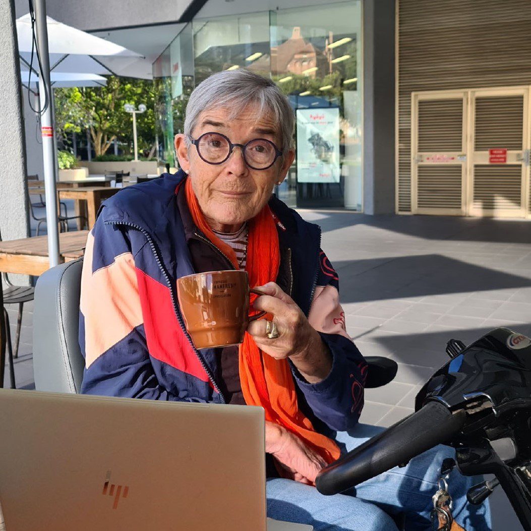 A lady sitting on an electric scooter is holding a cup of coffee with a laptop on the table in front of her.