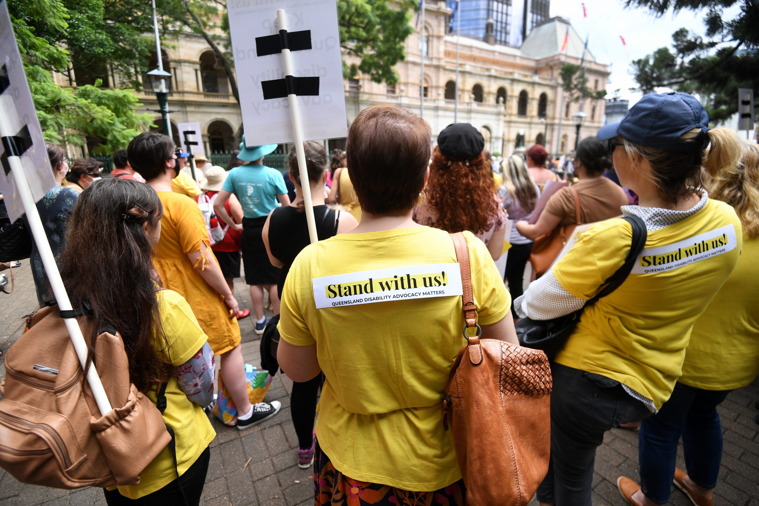 The backs of a group of people at the Stand with us! rally. Two people have stickers on their backs saying Stand with us!