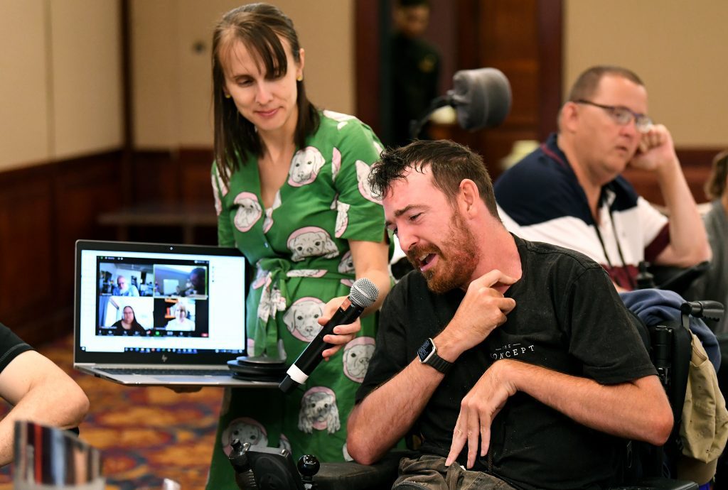A man in a black shirt, with brown hair and a short beard is sitting and talking into a microphone. A girl next to him in a green dress is holding the microphone and holding a laptop with people on zoom on the screen.
