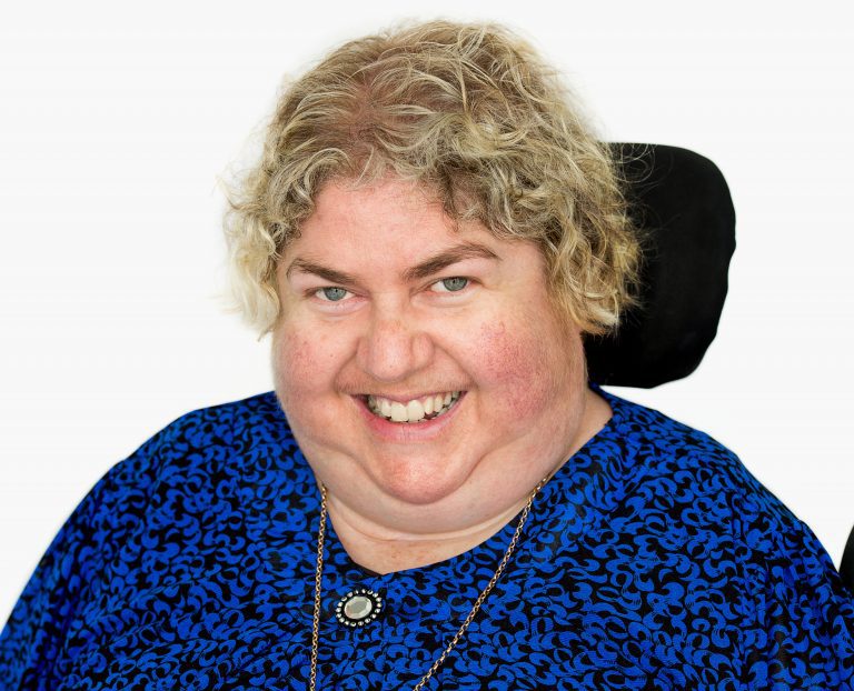 Q D N board member Sharon Boyce, in a blue dress, has short blonde hair and is smiling at the camera.