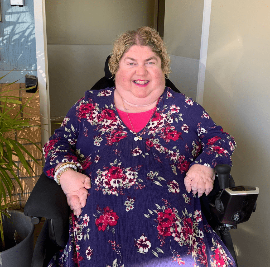 A lady with short blonde hair is in a wheelchair is wearing a blue dress with pink flowers is smiling at the camera.