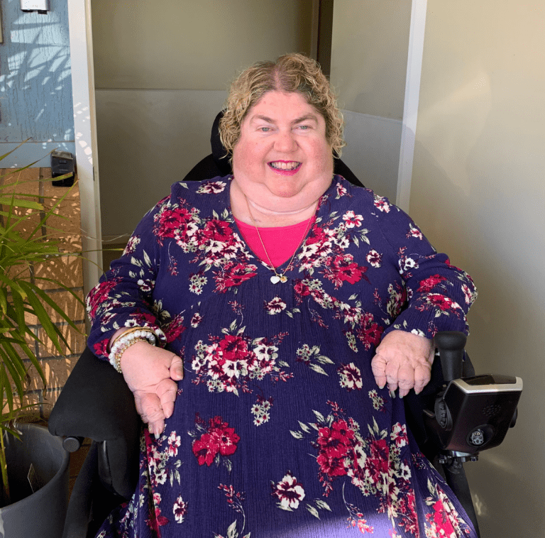 A lady in a blue dress with pink flowers in a wheel chair smiling at the camera.