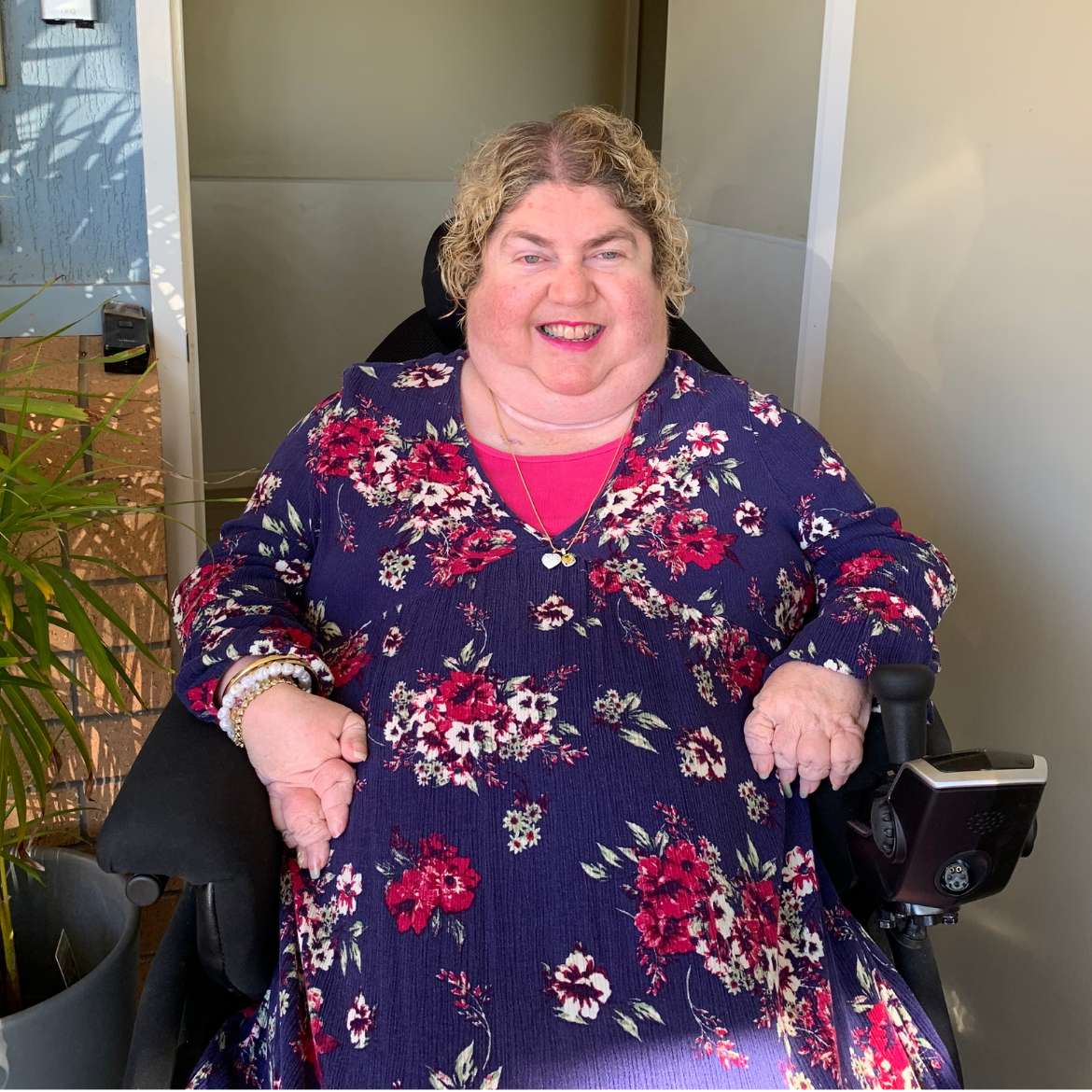 A lady in a blue dress with pink flowers in a wheel chair smiling at the camera.
