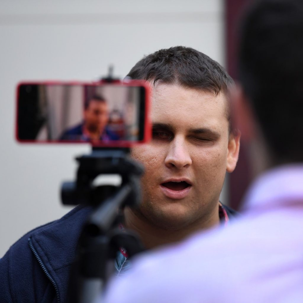 A man with his back to the camera is sitting behind a phone on a tripod. The man and the phone are facing facing a man is being filmed and is looking at the camera.