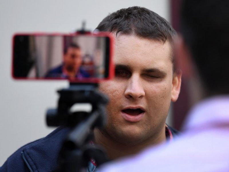 A man with his back to the camera is sitting behind a phone on a tripod. The man and the phone are facing facing a man is being filmed and is looking at the camera.