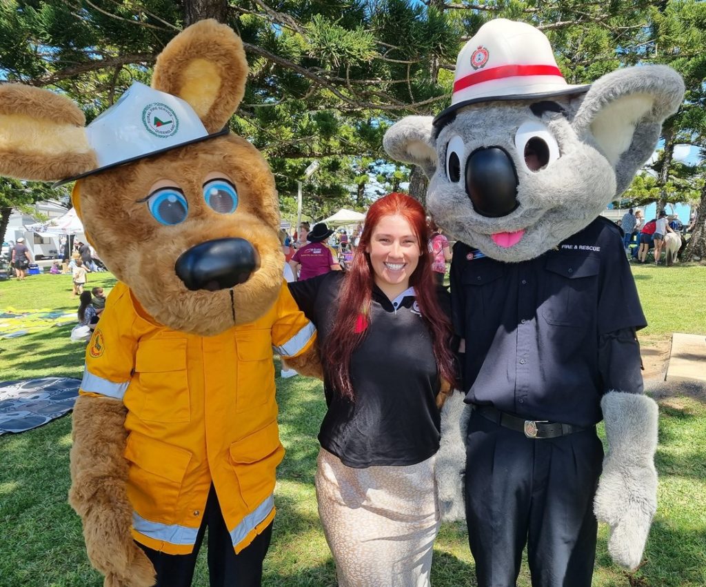 Lisa, a QDN staff member with long red hair and wearing a blue polo, is standing with her arms around two people in large custumes portraying Bushy the Kangaroo, and Blazer the Koala. They are the Queensland Fire and Emergency Services mascots.