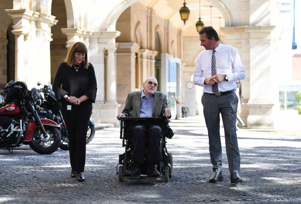 Three people outside walking. There is a lady wearing black, a man in a blazer in a wheelchair and another man on the other side wearing a white shirt and tie.