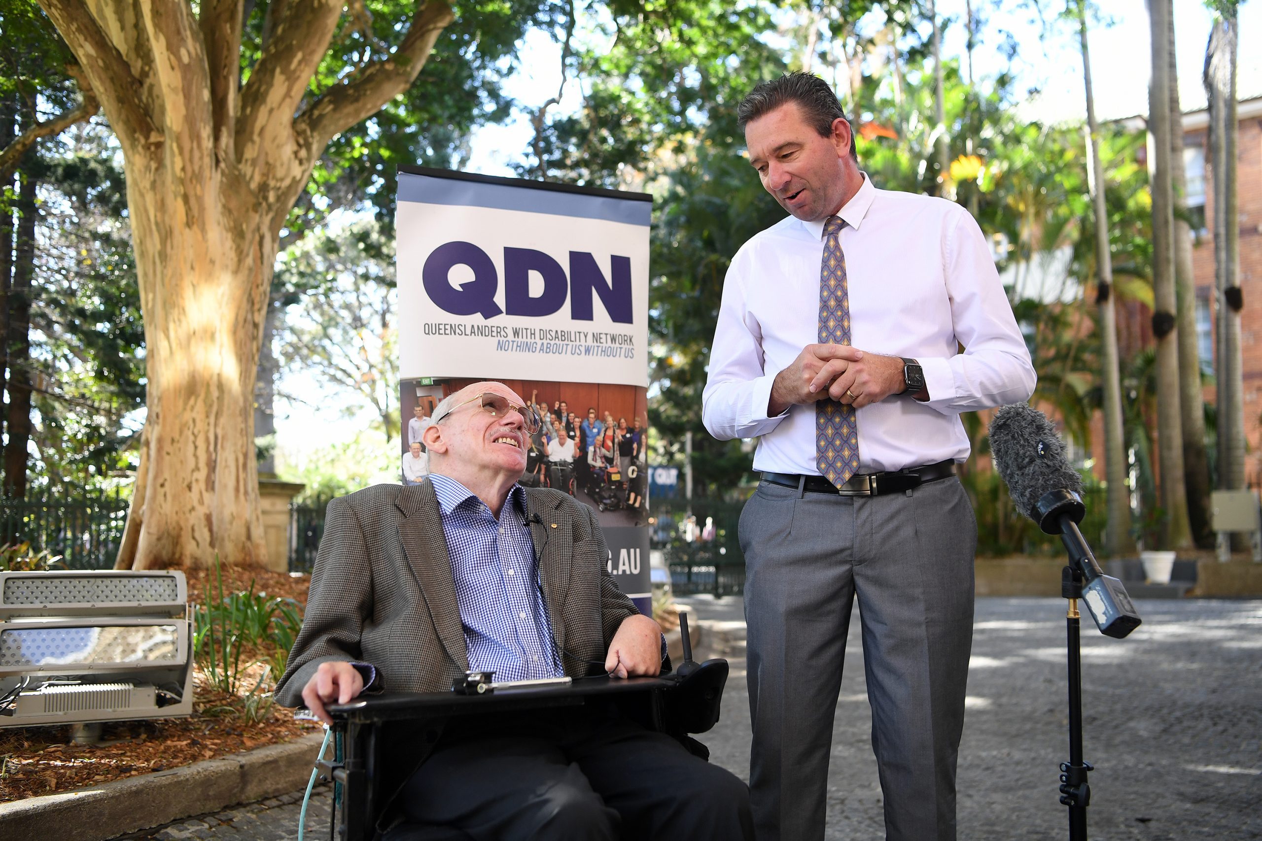 Two men outside under some trees. A man is in a wheelchair looking up at another man who is standing. There is a microphone in the foreground.