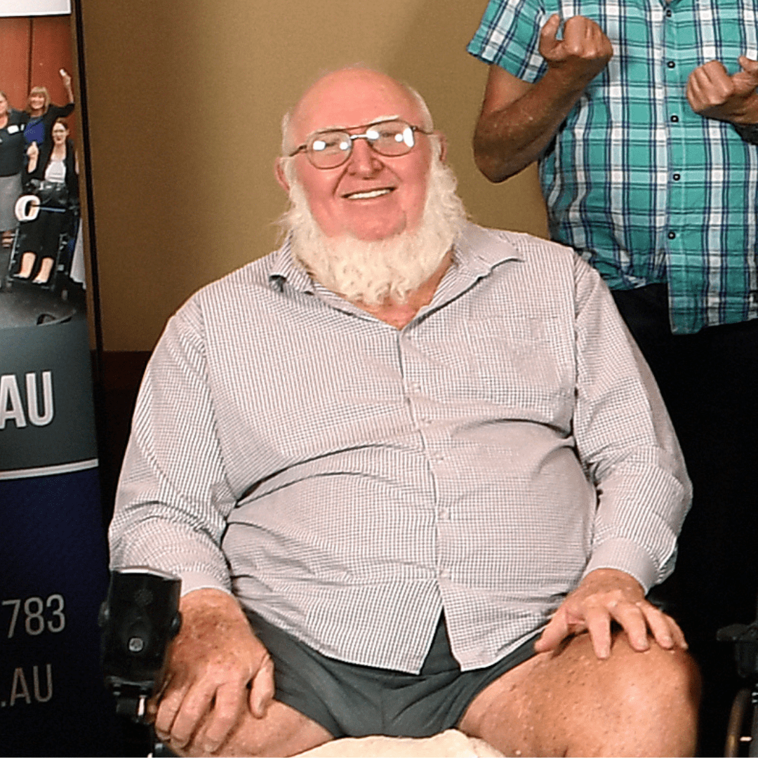 A man with a white beard, with glasses and wearing a long sleeved button up shirt. He is sitting in a wheelchair.