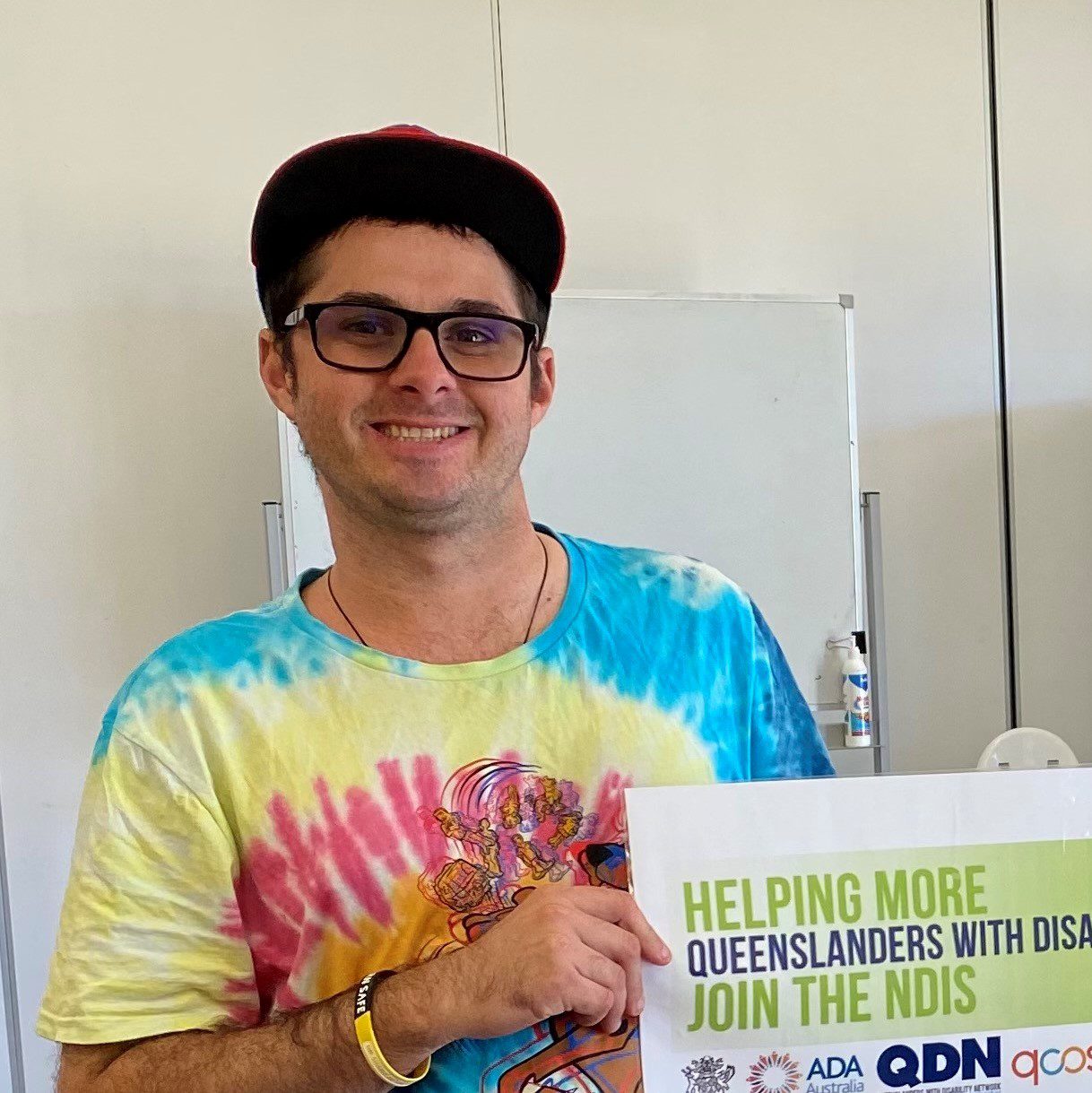 A man wearing a cap and glasses and a tie dyed bright shirt smiling at the camera.