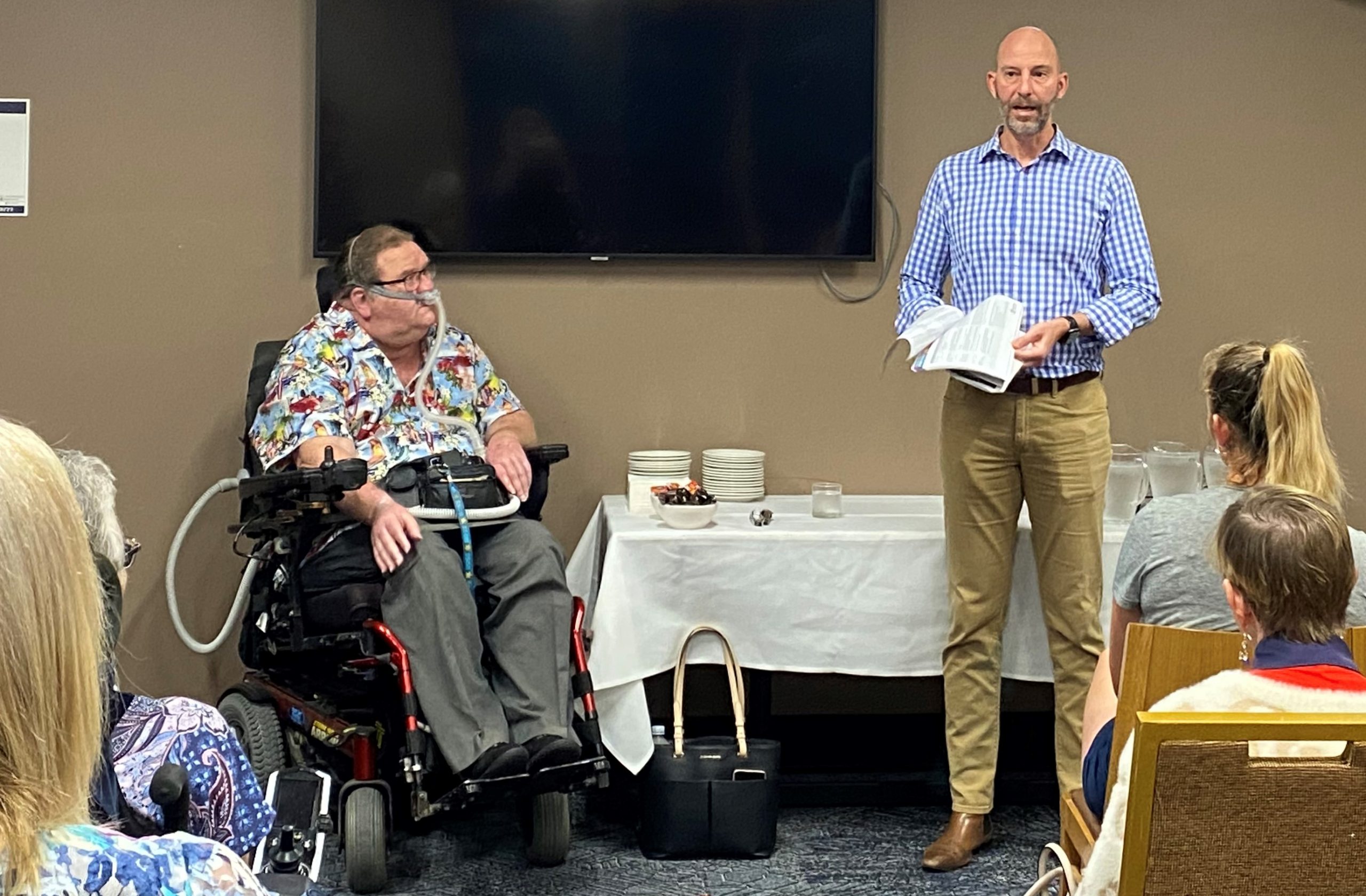 A man standing at the front of the room presenting. He is wearing a light blue long sleeved button up shirt and beige long pants. There is a man sitting in a wheelchair next to him. 