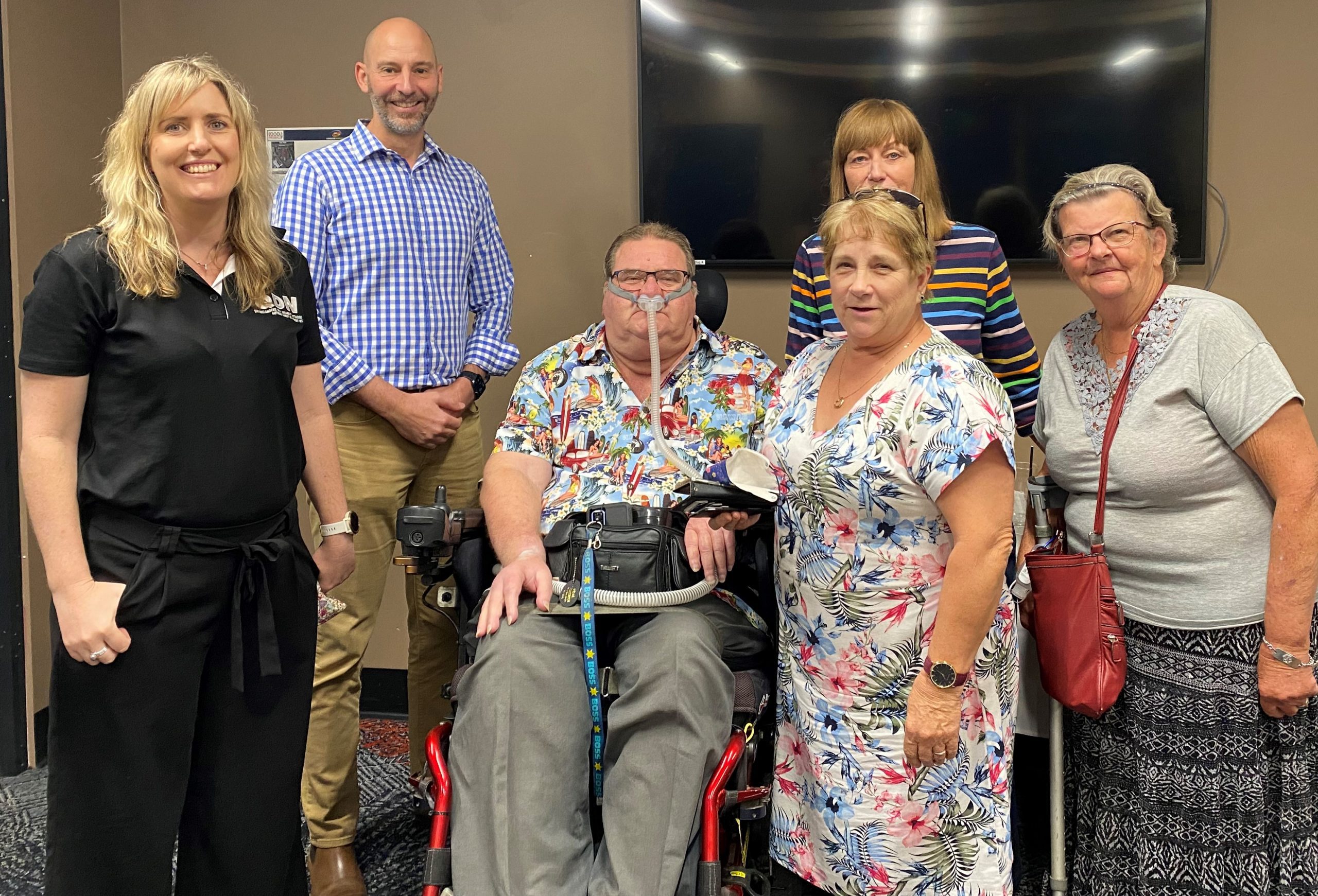 A group of 5 people standing and 1 sitting in a wheelchair. They are all looking at the camera smiling. 