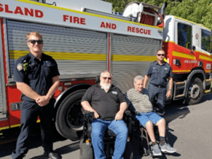 Picture of Queensland Fire and Rescue staff, beside a fire truck in Logan and two QDN members with them sitting in wheelchairs..