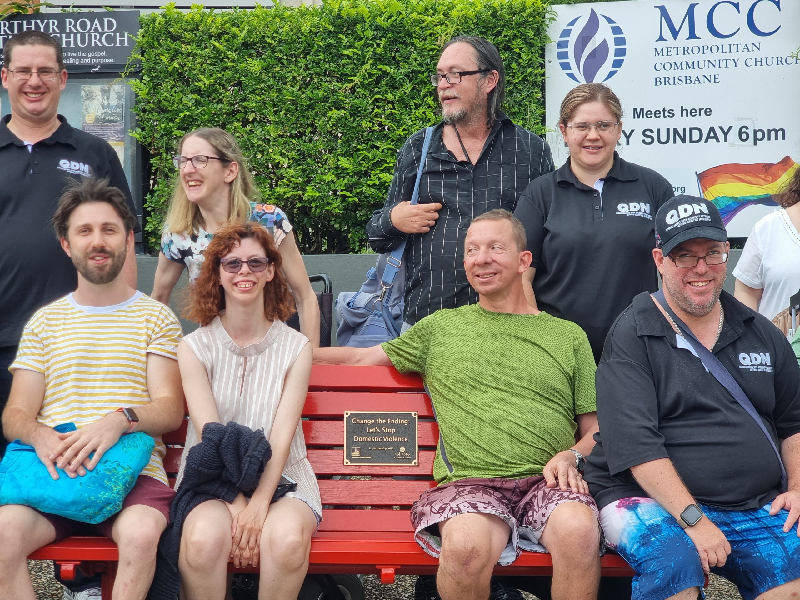 A group of people sitting on a bench and some standing behind. They are all smiling and most are looking at the camera.
