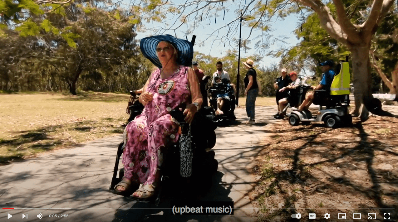 Screen shot of a video. There is a woman in a wheel chair going down a path in a park.