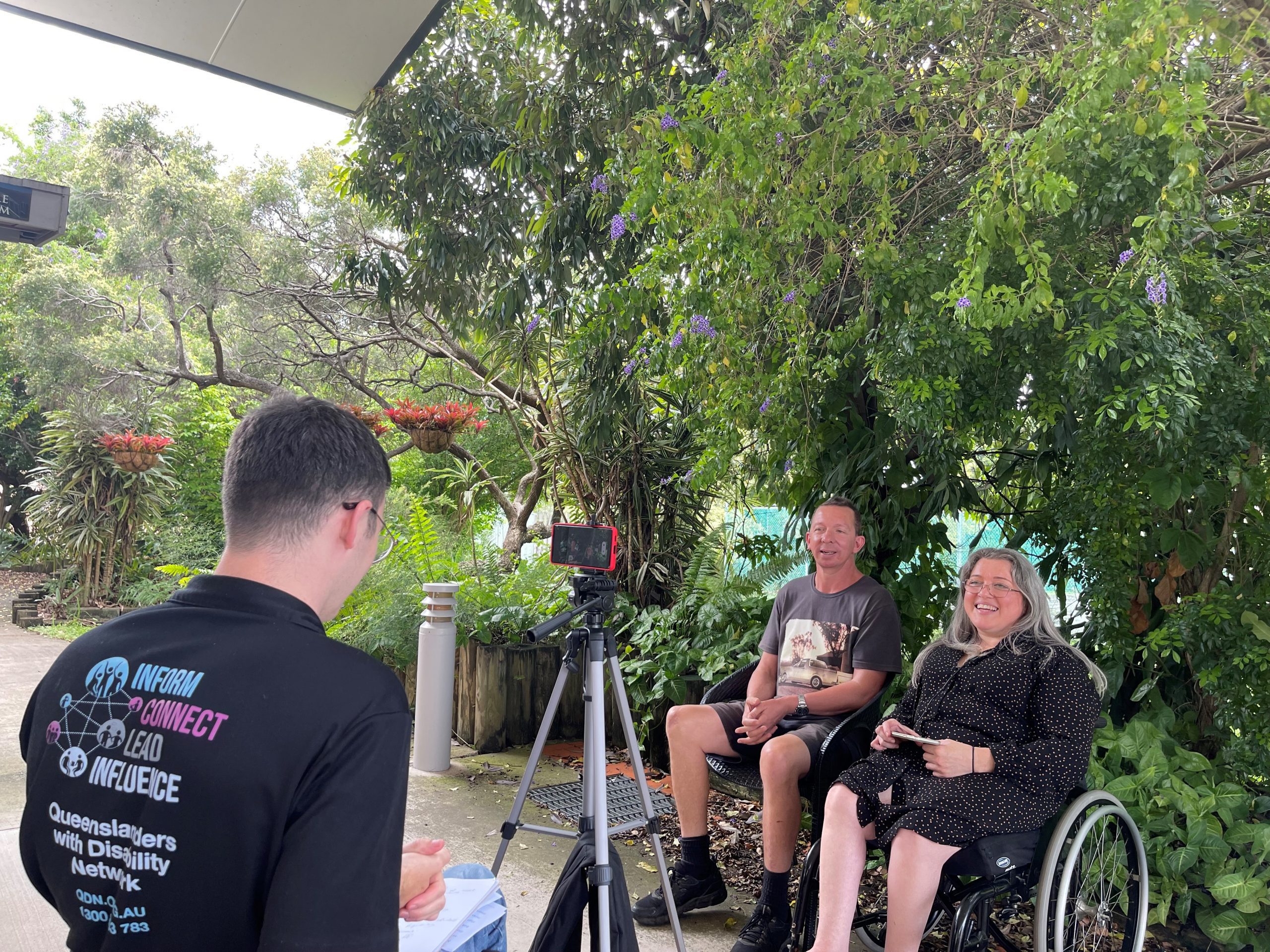 A woman in a wheelchair with long grey hair and a man sitting next to her with short brown hair. They are looking into a phone on a tripod with a man with his back to us wearing a QDN polo shirt.