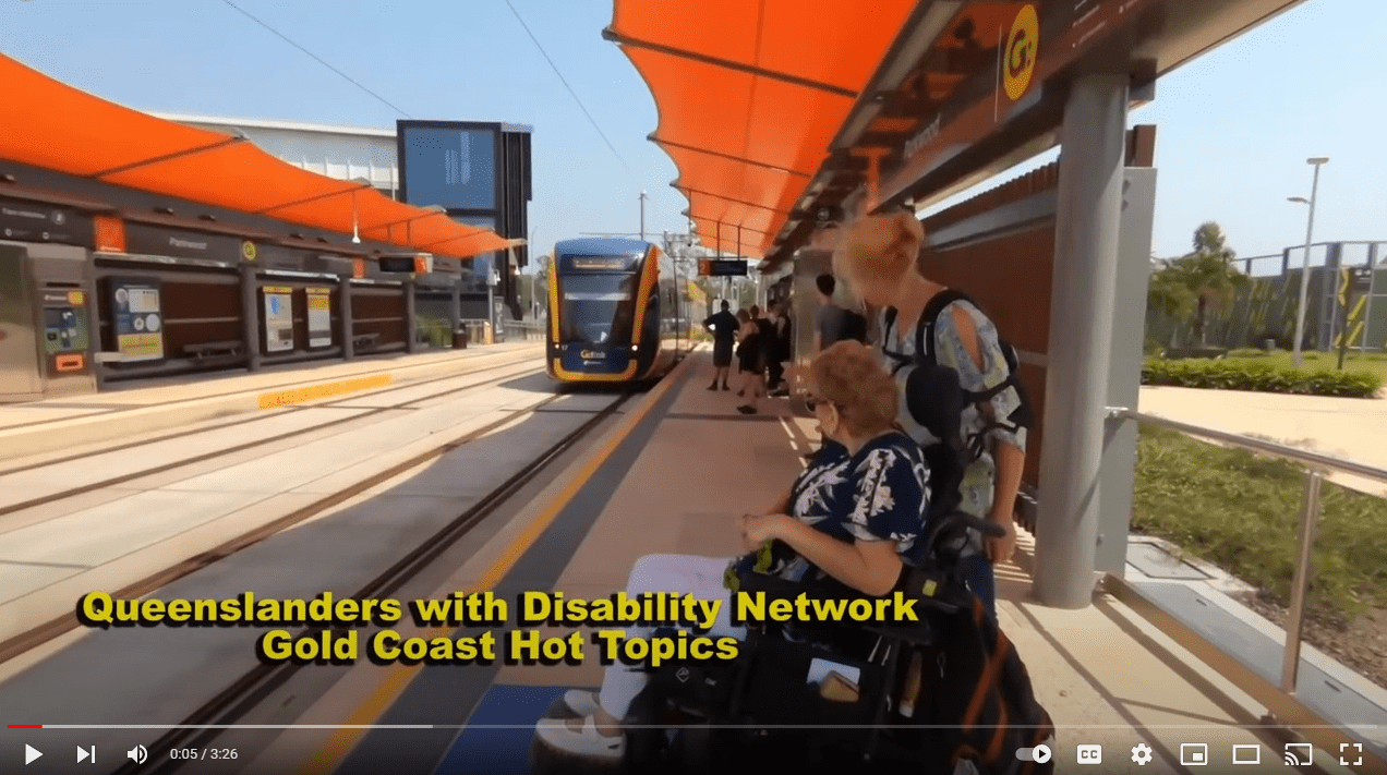Screen shot of the beginning of a video. There are two women, one in a wheelchair, waiting for a tram. The text says Queenslanders with Disability Network Gold Coast Hot Topics.