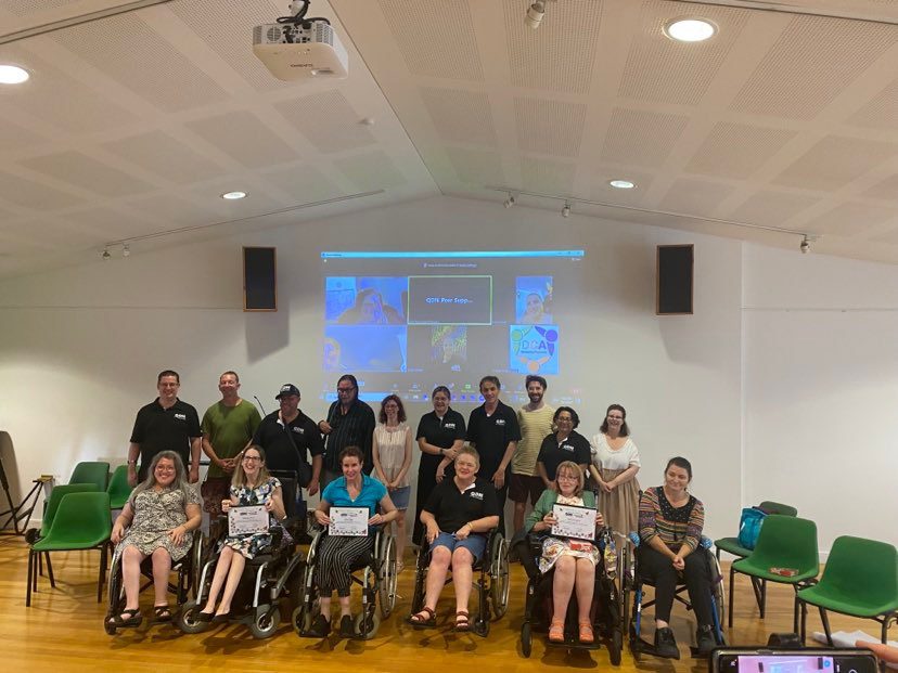 A group of people, the first row is all people sitting in wheelchairs, then a row of people standing behind them. There is a projector on the wall of people in a zoom call.