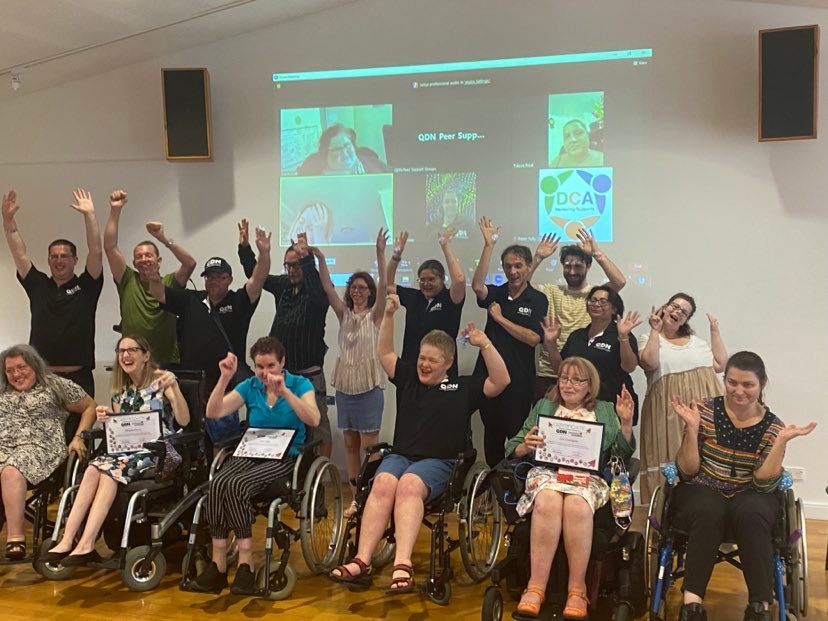 A group of people, the first row is all people sitting in wheelchairs, then a row of people standing behind them. There is a projector on the wall of people in a zoom call. Some are holding signs and others have there hands in the air cheering.