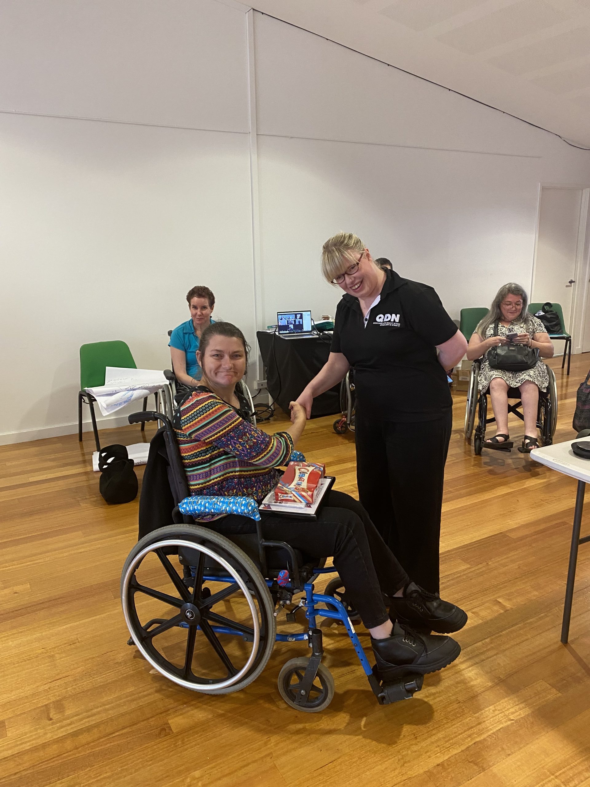 A woman in a striped colourful shirt, sitting in a wheel chair shaking hands with a lady standing wearing a QDN shirt. They are looking at the camera smiling.
