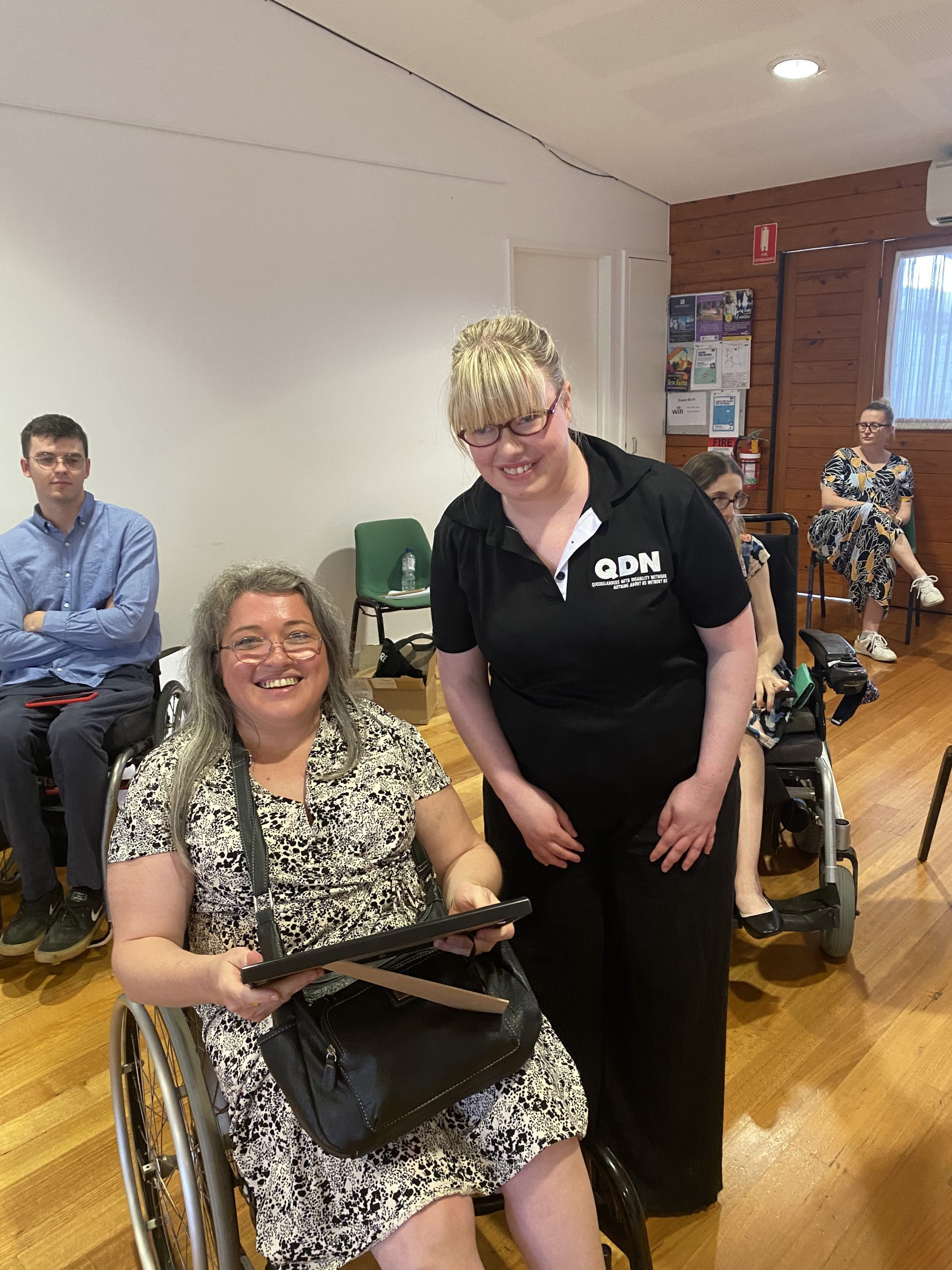 A woman sitting a wheelchair and another woman standing next to her. They are both smiling into the camera.