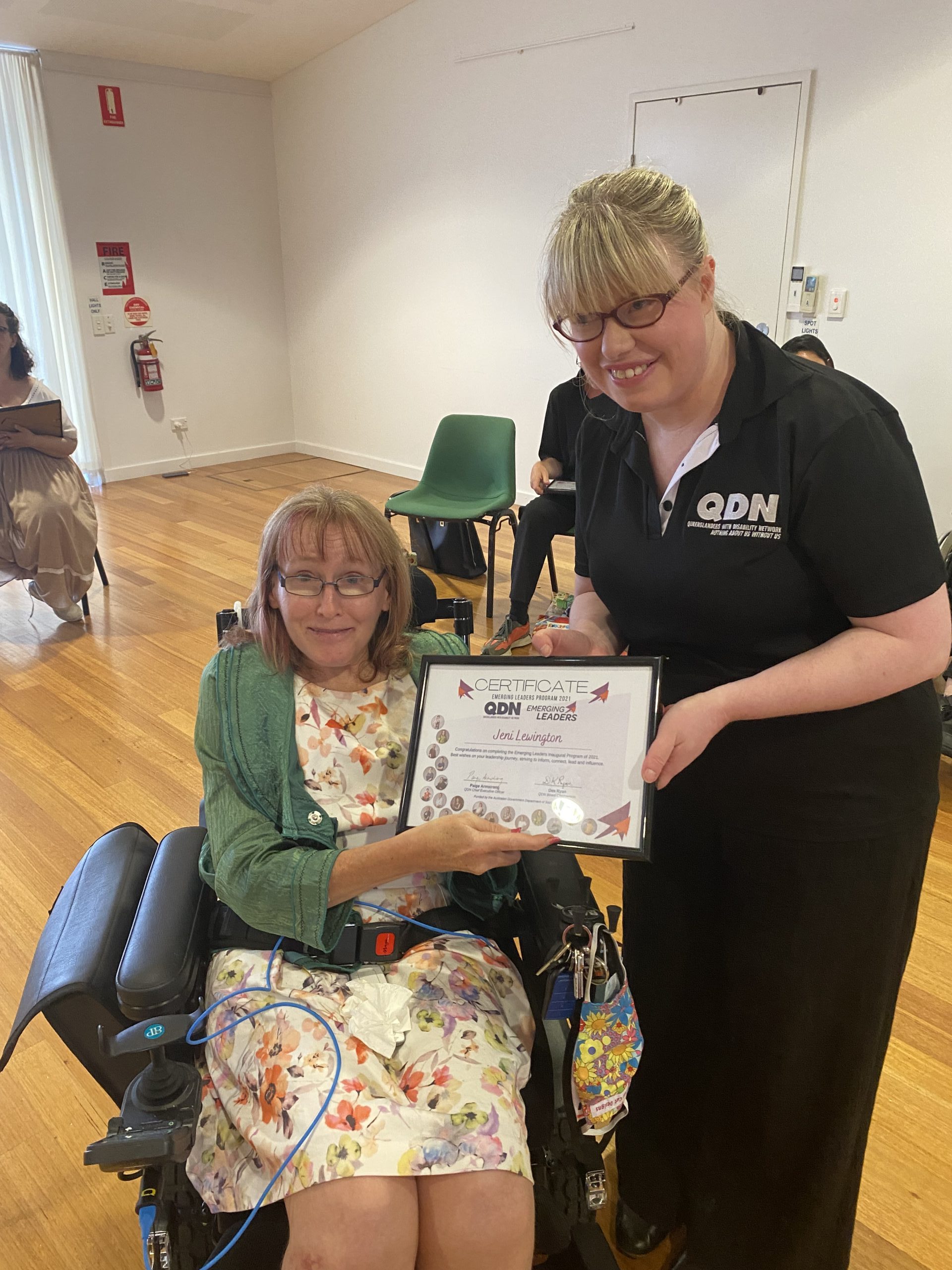 A lady in a wheel chair and another lady standing next to her wearing a QDN shirt. They are holding a framed certificate together and looking at the camera smiling.
