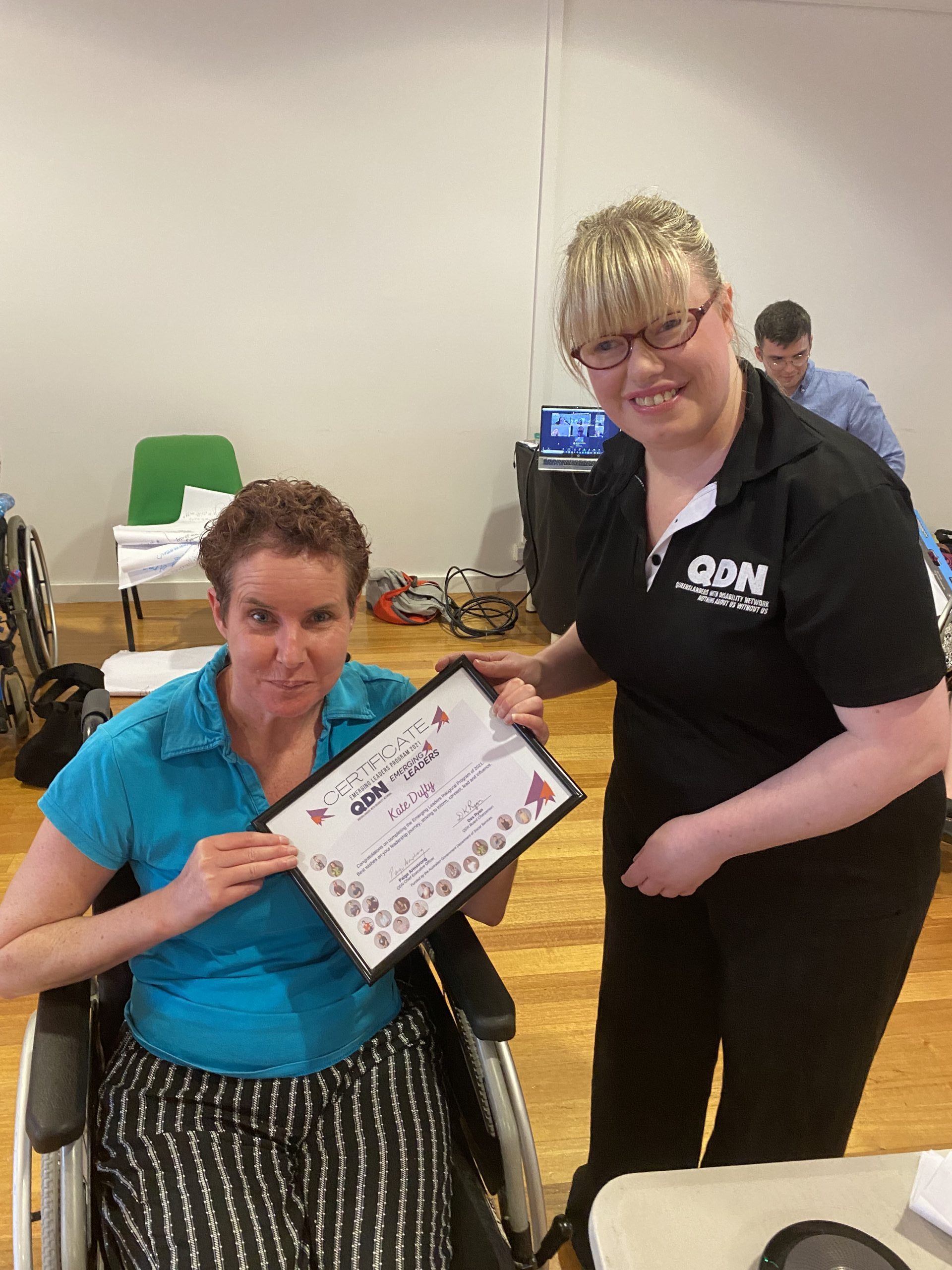 A lady wearing a bright blue shirt sitting in a wheelchair and another lady in a QDN shirt standing next to her handing her a framed certificate. They are both looking at the camera smiling.