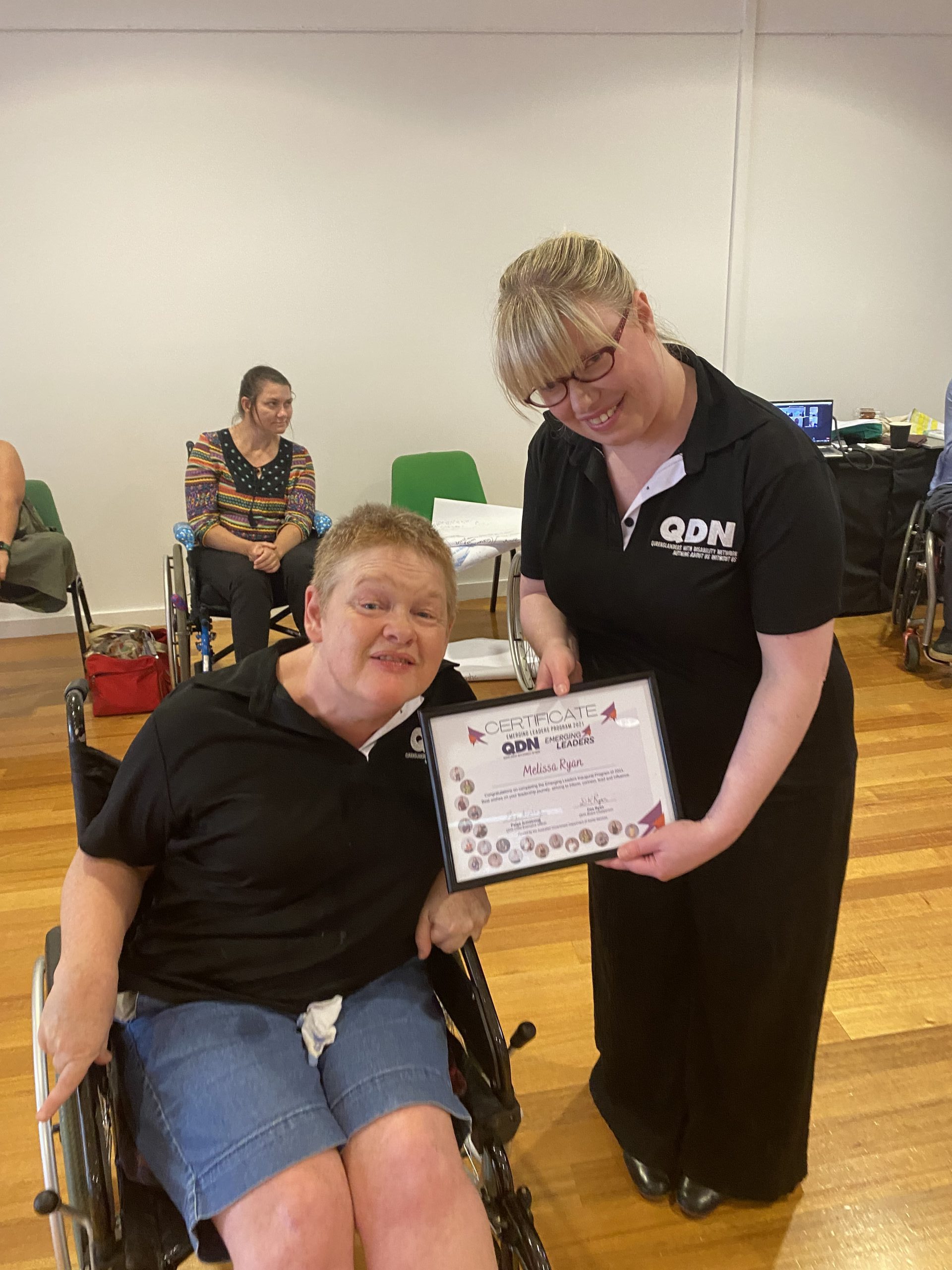 Two ladies in QDN shirts, one in a wheelchair and one standing. They are holding a framed certificate and looking at the camera smiling.
