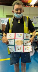 A man with brown hair, wearing a face mask and a high vis vest over a black shirt. He is holding up a resource around disaster emergency planning.