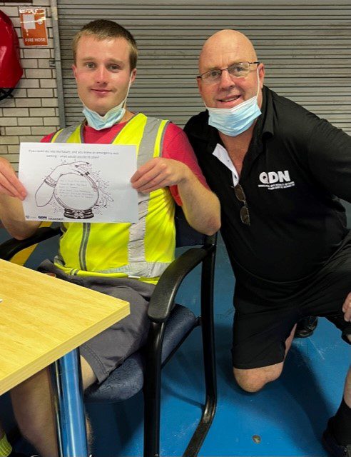 A man sitting in a chair with a red shirt and a yellow high vis vest, he is holding up a resource. There is a man squatting behind him wearing a QDN polo and smiling at the camera.