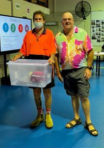 Two men standing next to each other. One is wearing a bright orange polo and is holding a large plastic container. The man on his right is wearing a multicoloured shirt and glasses.