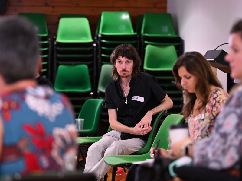 People sitting on chairs in a circle, the photo focuses on one man with brown hair wearing a black polo.