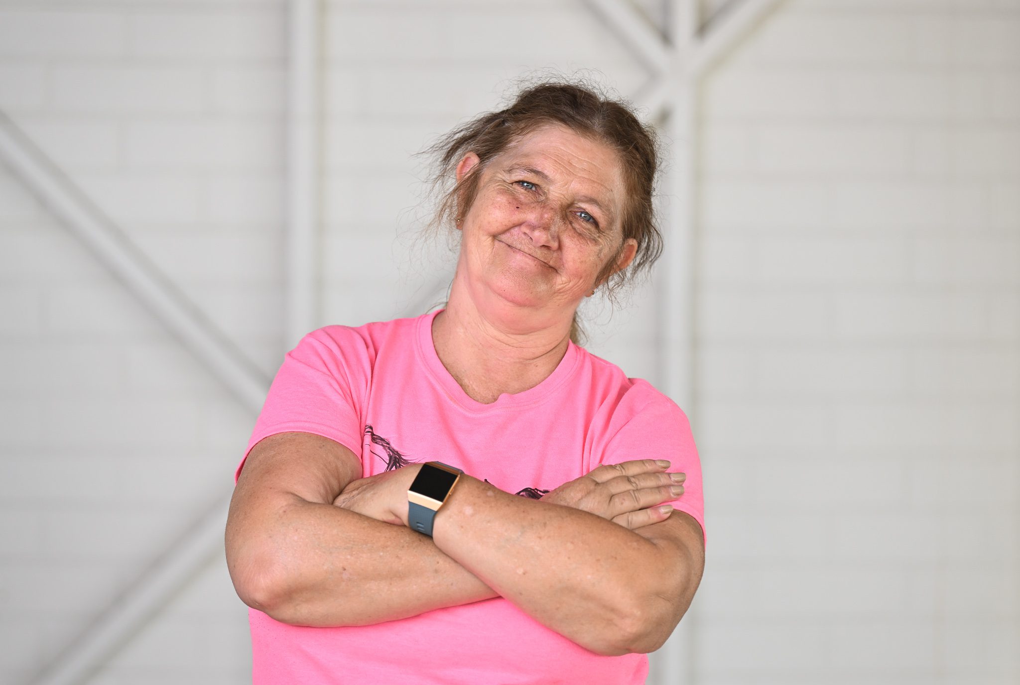 A lady wearing a pink tshirt, with her arms crossed looking at the camera smiling.