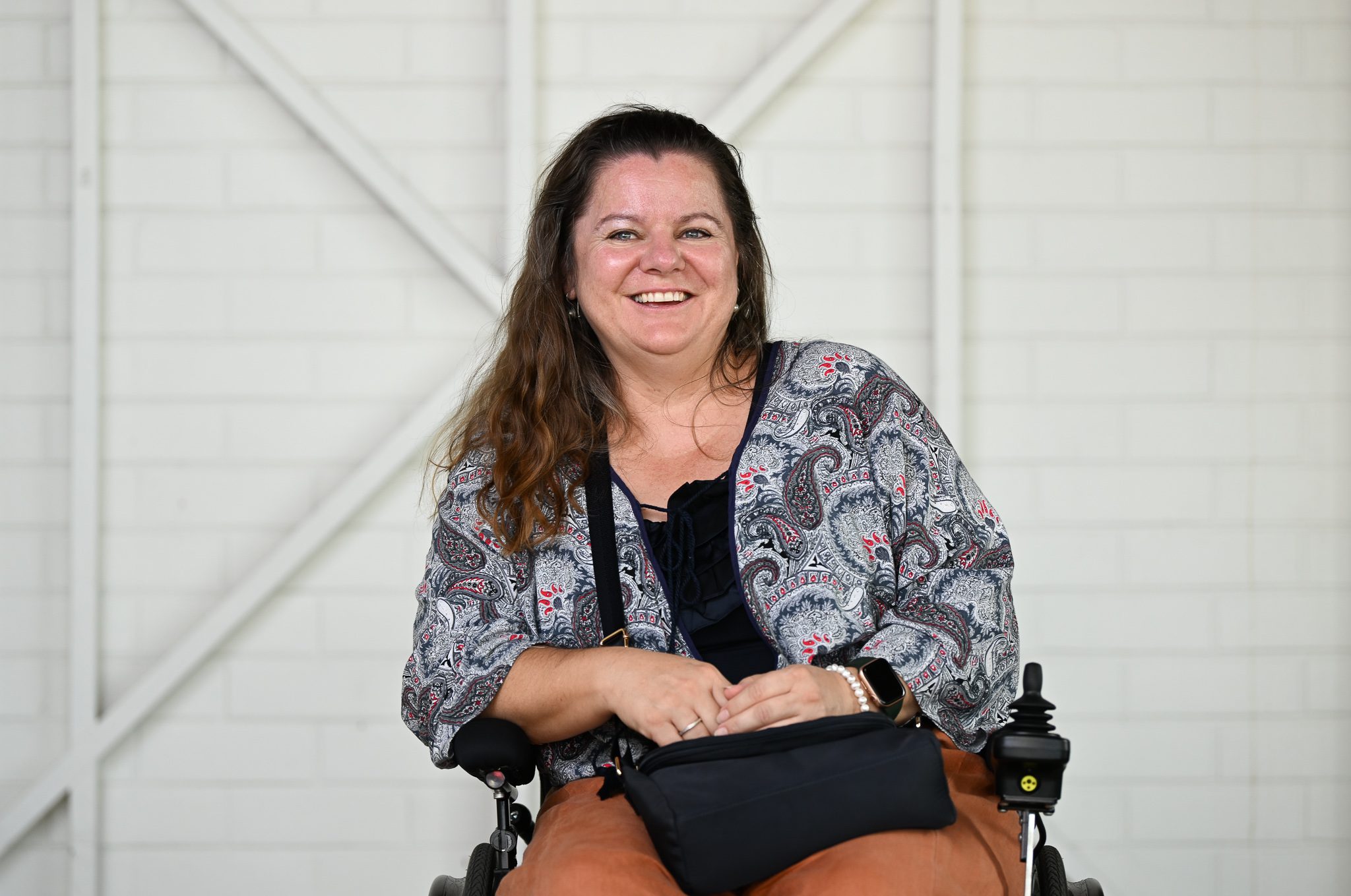 A woman with long brown hair, wearing a patterened shirt and holding a black back, she is sitting in a wheelchair and smiling at the camera.