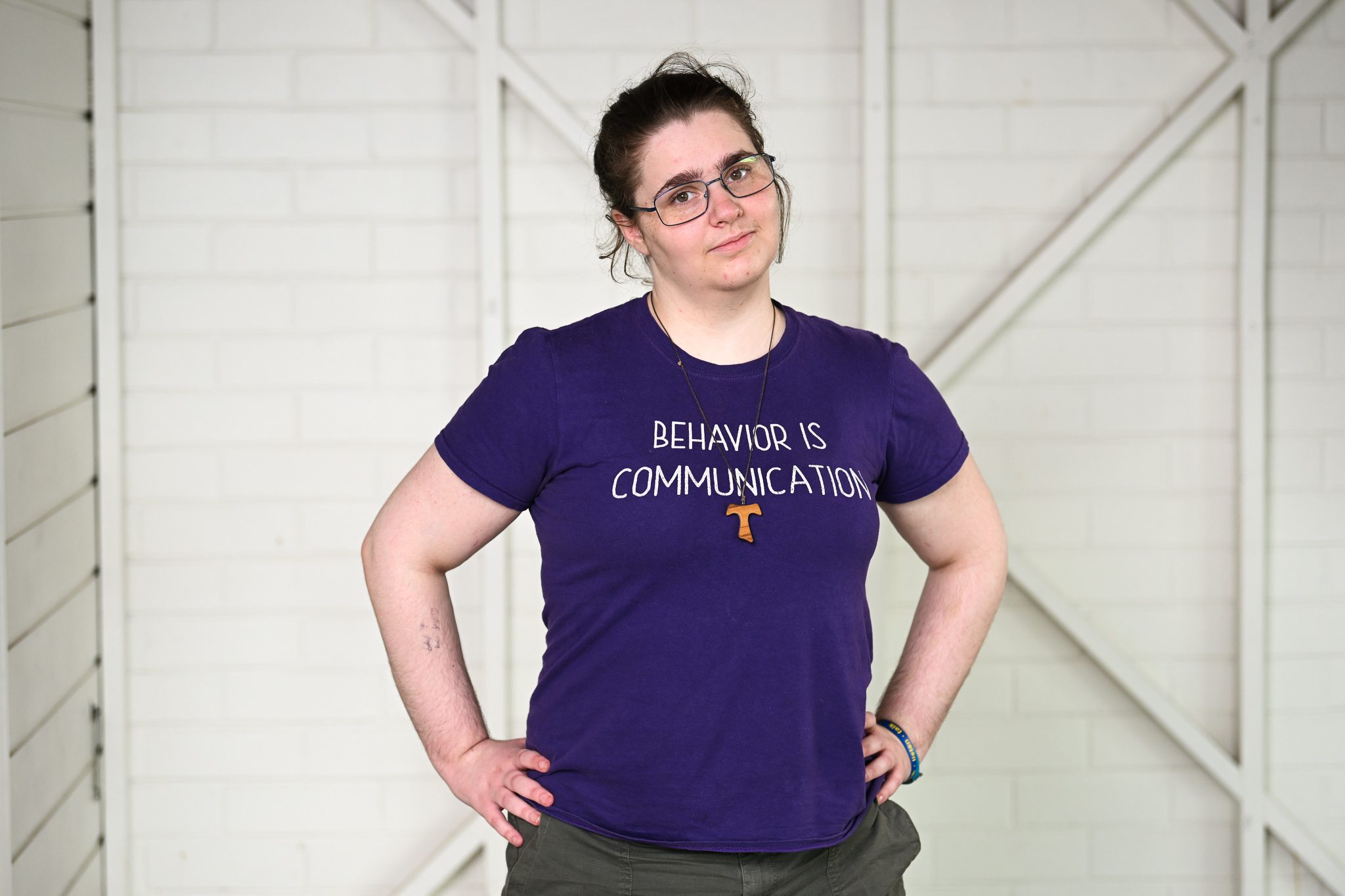 A girl with brown hair and glasses, wearing a purple tshirt, she has her hands on her hips and smiling at the camera.