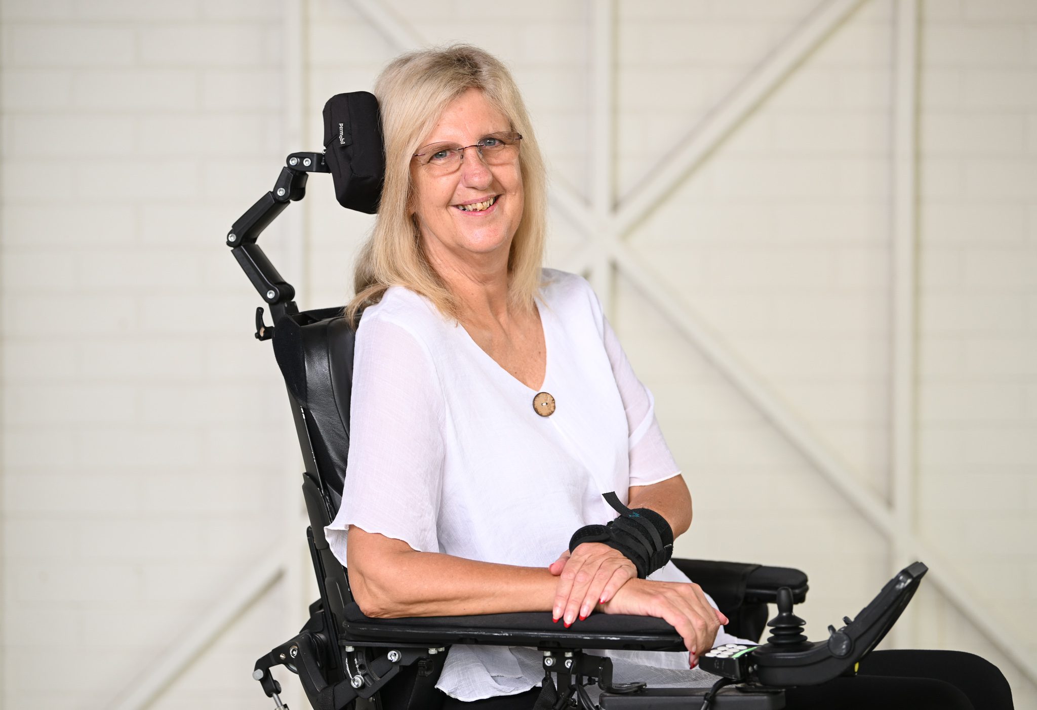 A woman with long blonde hair, wearing a white blouse, sitting in a wheelchair and looking at the camera smiling.