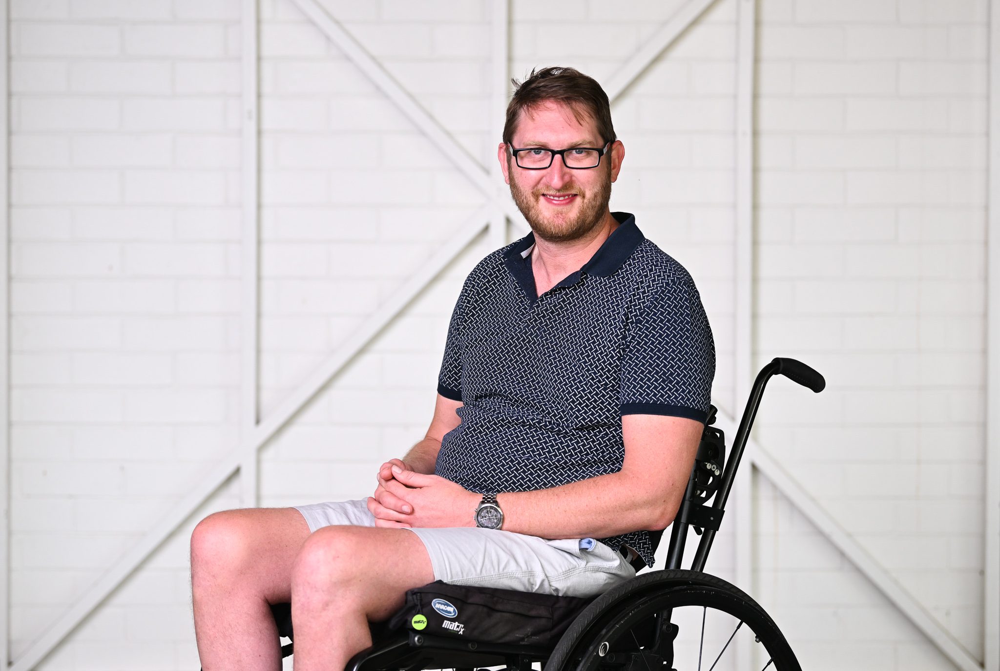 A man with short brown hair wearing a black polo, sitting in a wheelchair and smiling at the camera.