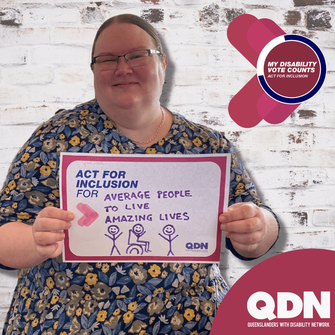 A woman wearing a floral dress, with her hair pulled back and wearing glasses. She is smiling at the camera and holding a sign that says Act for inclusion for and written in pen "Average people to live amazing lives". QDN Queenslanders with Disability Network.
