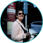 There is a photo in a circle with a woman wearing dark glasses and holding a cane, standing next to a taxi sign.