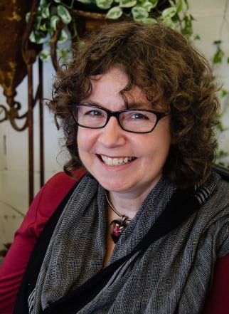 A woman with brown curly hair, wearing glasses and a gray scarf. Looking at the camera smiling. 