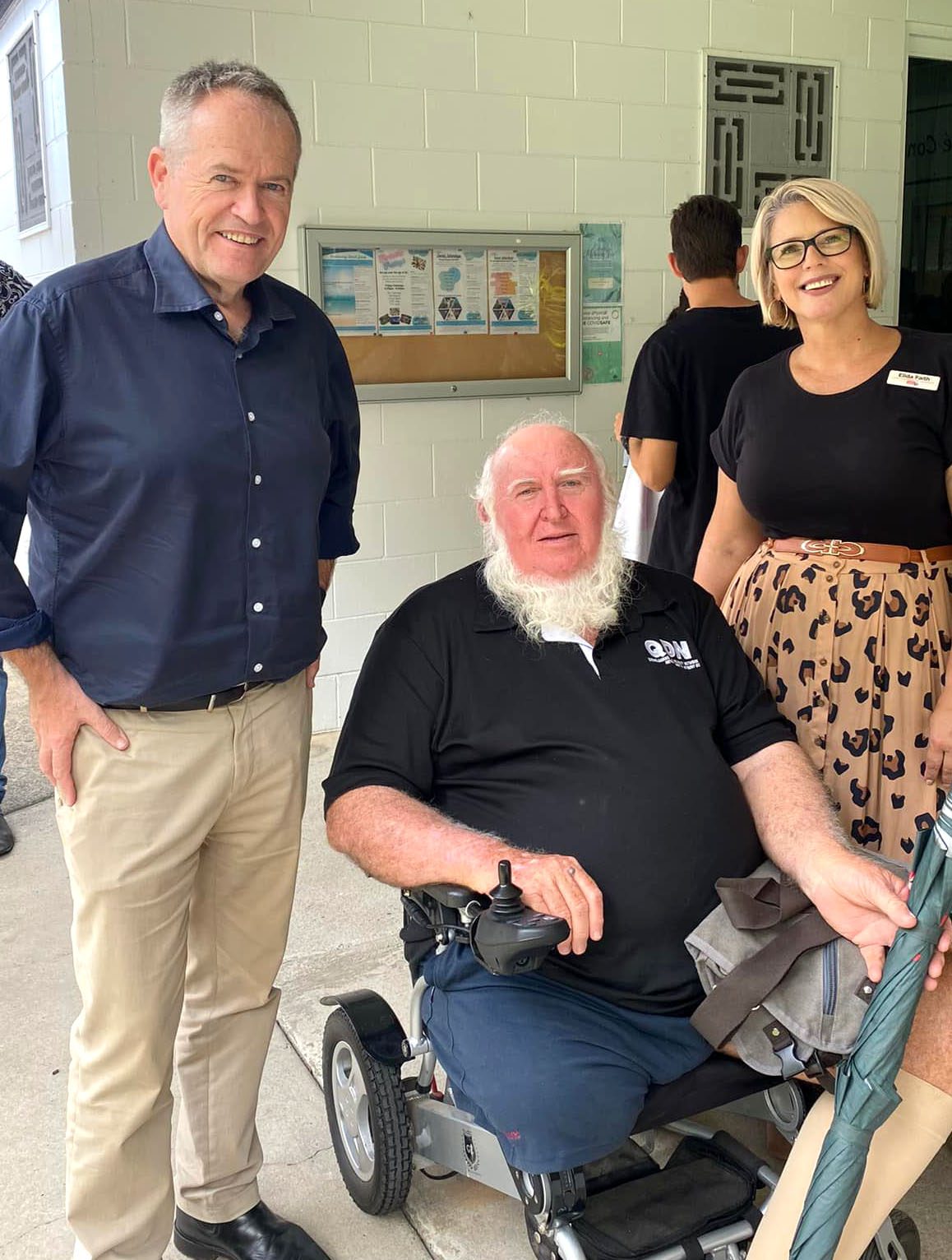 Three people smiling at the camera. One man standing wearing a blue long sleeved business shirt, next to him a man is sitting in a wheelchair wearing a QDN black polo, next to him is a woman standing wearing a black shirt and floral skirt. 