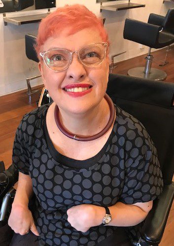 A woman with short light pink hair, wearing glasses and looking up smiling at the camera.