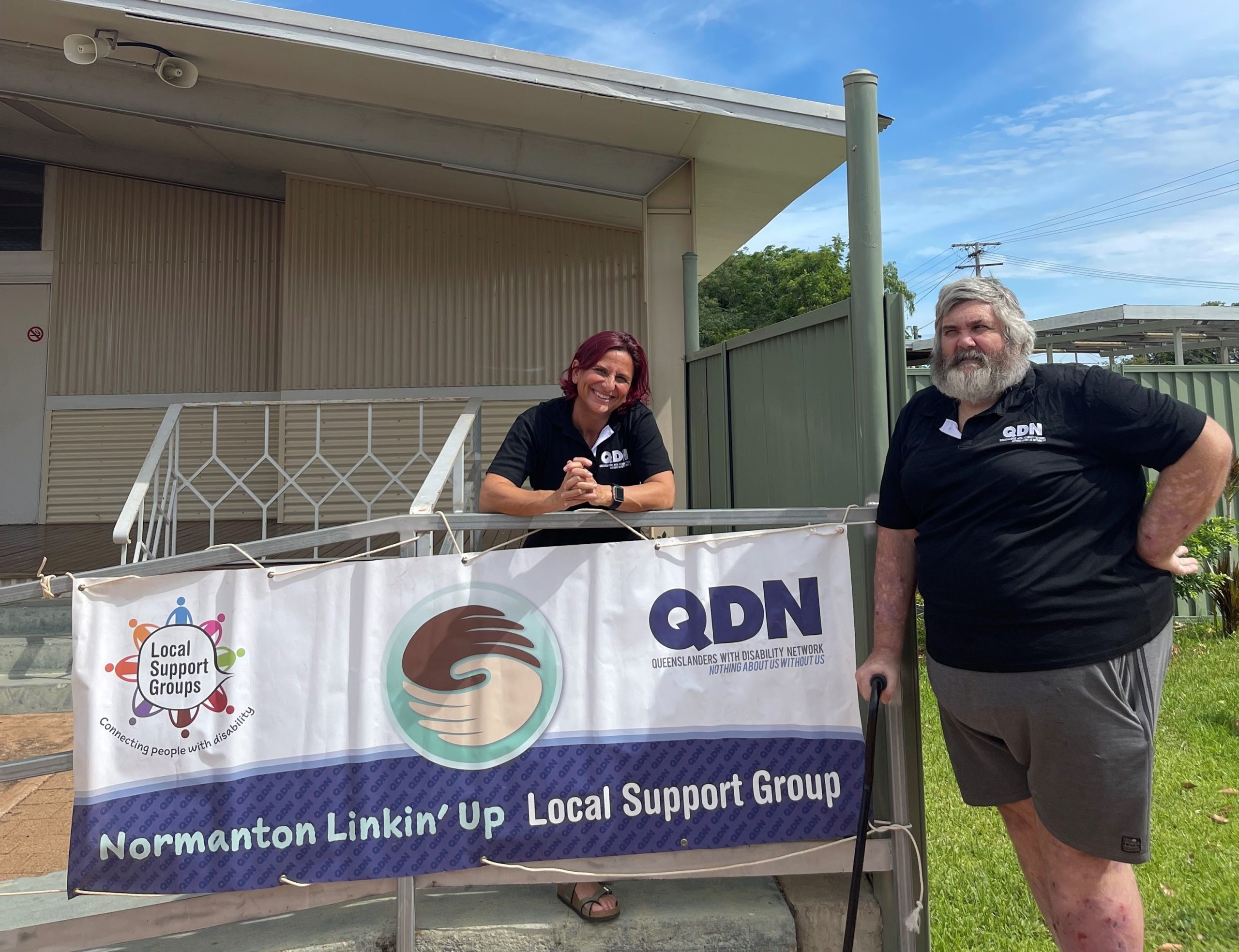 A man and woman both wearing black QDN polos standing next to a large sign that says Normanton Linkin' Up Local Support Group. QDN Queenslanders with Disability Network. Nothing about us without us. 